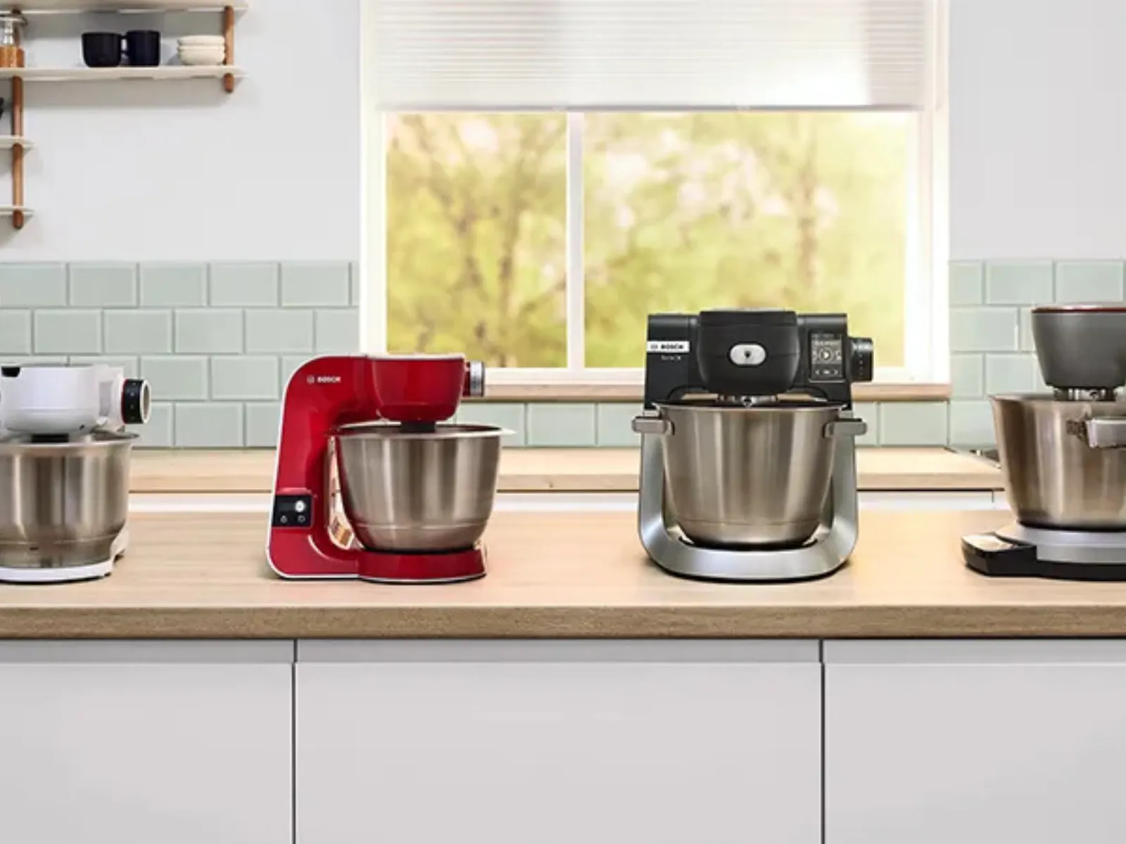 Four colorful stand mixers in a row on a kitchen counter, window view of greenery behind them.