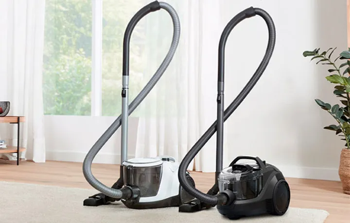 Two Bosch canister vacuum cleaners, one white and one black, stand side-by-side on a light rug in a bright room with a large window.