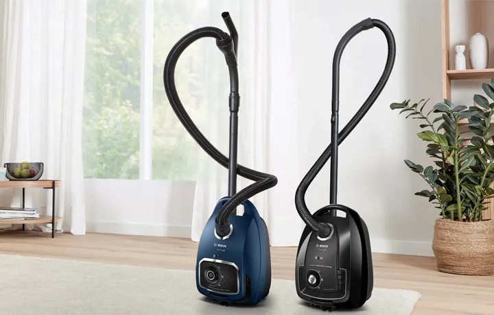 Two Bosch canister vacuum cleaners, one blue and one black, stand side-by-side on a light rug in a bright room with a large window.