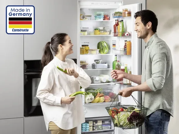 Frau mit Gemüse spricht mit Mann mit Obstkorb neben vollem Kühlschrank.