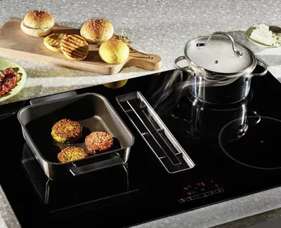 Kombi-Induktionskochfeld mit Dunstabzug. Auf dem Kochfeld befinden sich ein Topf und eine Pfanne mit Bratlingen.