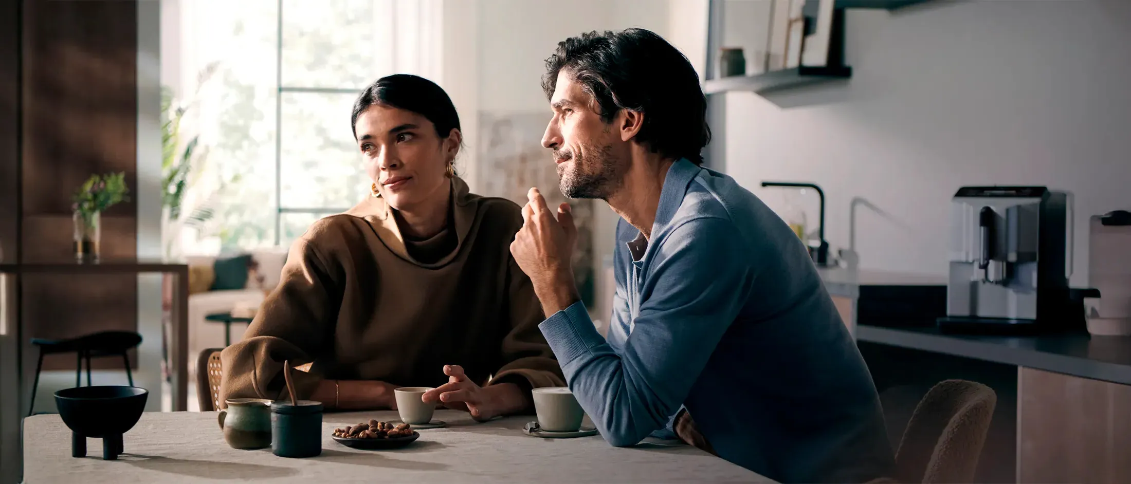 שני אנשים נהנים מקפה יחד במטבח מודרני, עם מכונת קפה אוטומטית מלאה של סימנס ברקע.