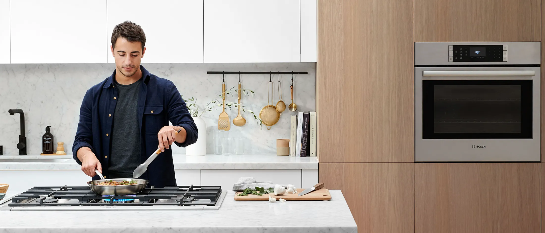 A person stands at a modern kitchen counter, cooking in a pan on a gas stove, surrounded by utensils and ingredients.
