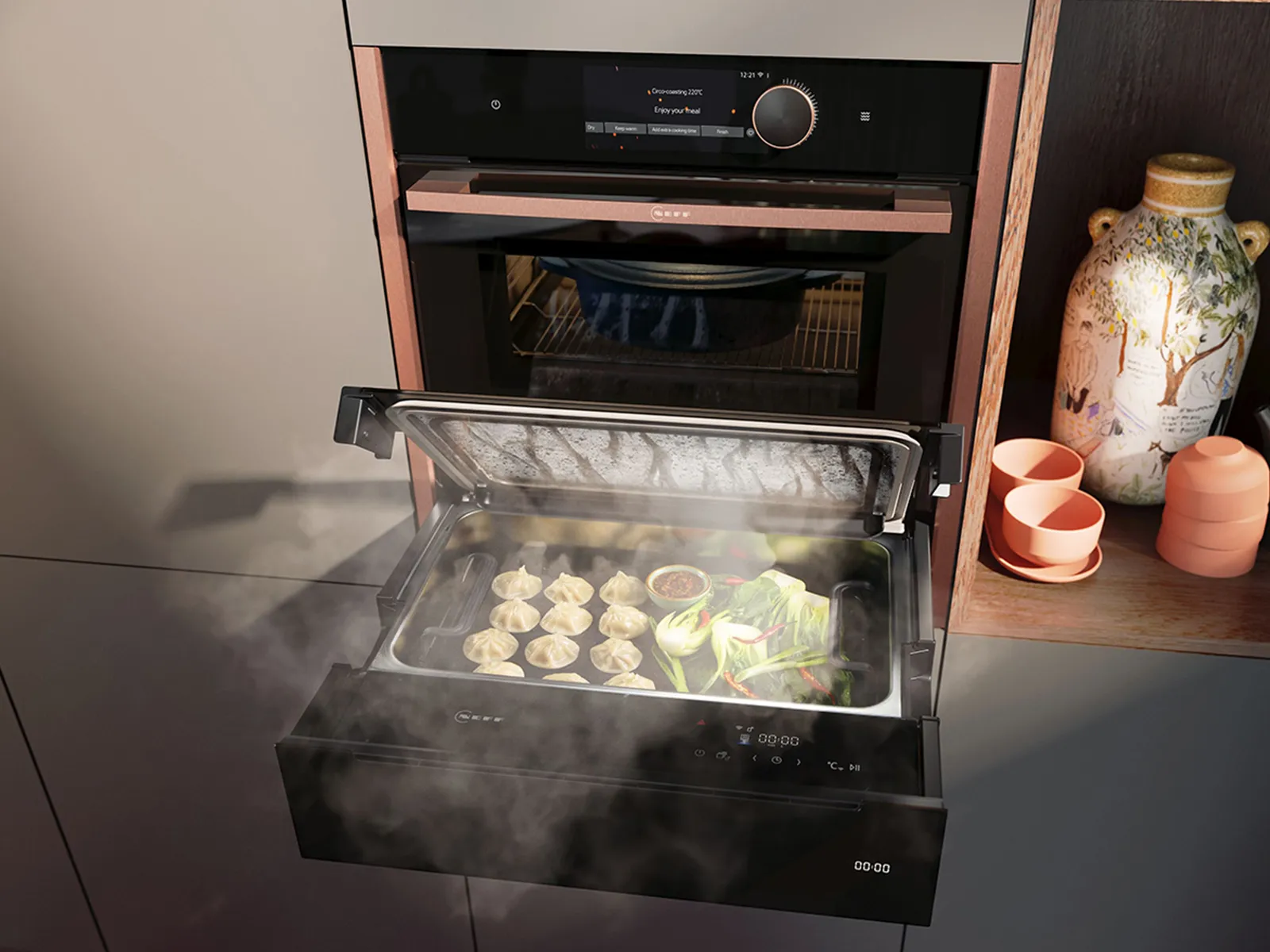A modern kitchen scene featuring a steam drawer filled with dumplings and vegetables, beside decorative ceramics and books.