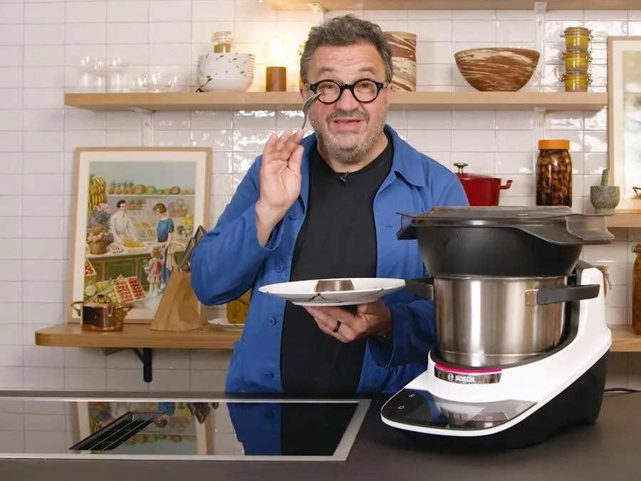 Yves Camdeborde debout dans une cuisine, tenant une assiette, avec un appareil Cookit sur le comptoir à côté de lui.