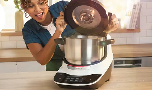 Una persona abre un Bosch Cookit con una olla metálica brillante, ubicada sobre una encimera de madera en una cocina luminosa.