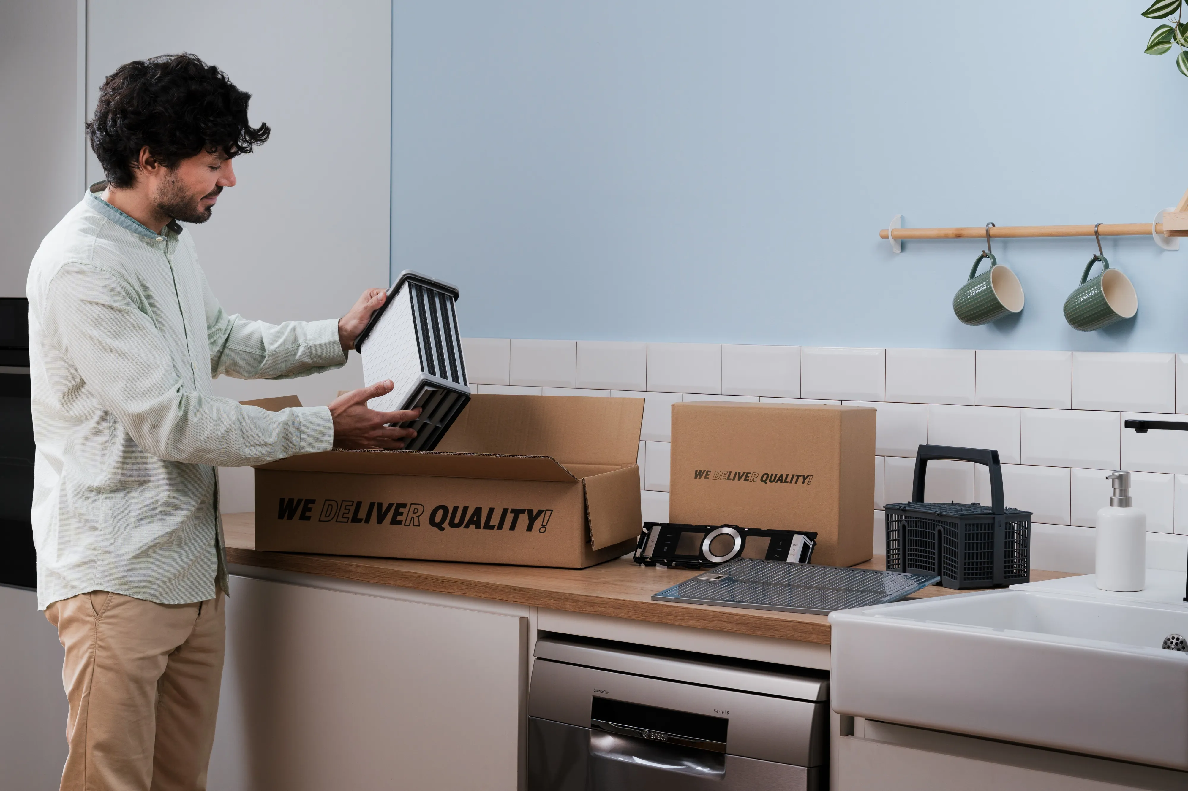 Homme debout dans une cuisine moderne ouvrant une boîte en carton marquée 'WE DELIVER QUALITY' contenant un panier en plastique noir