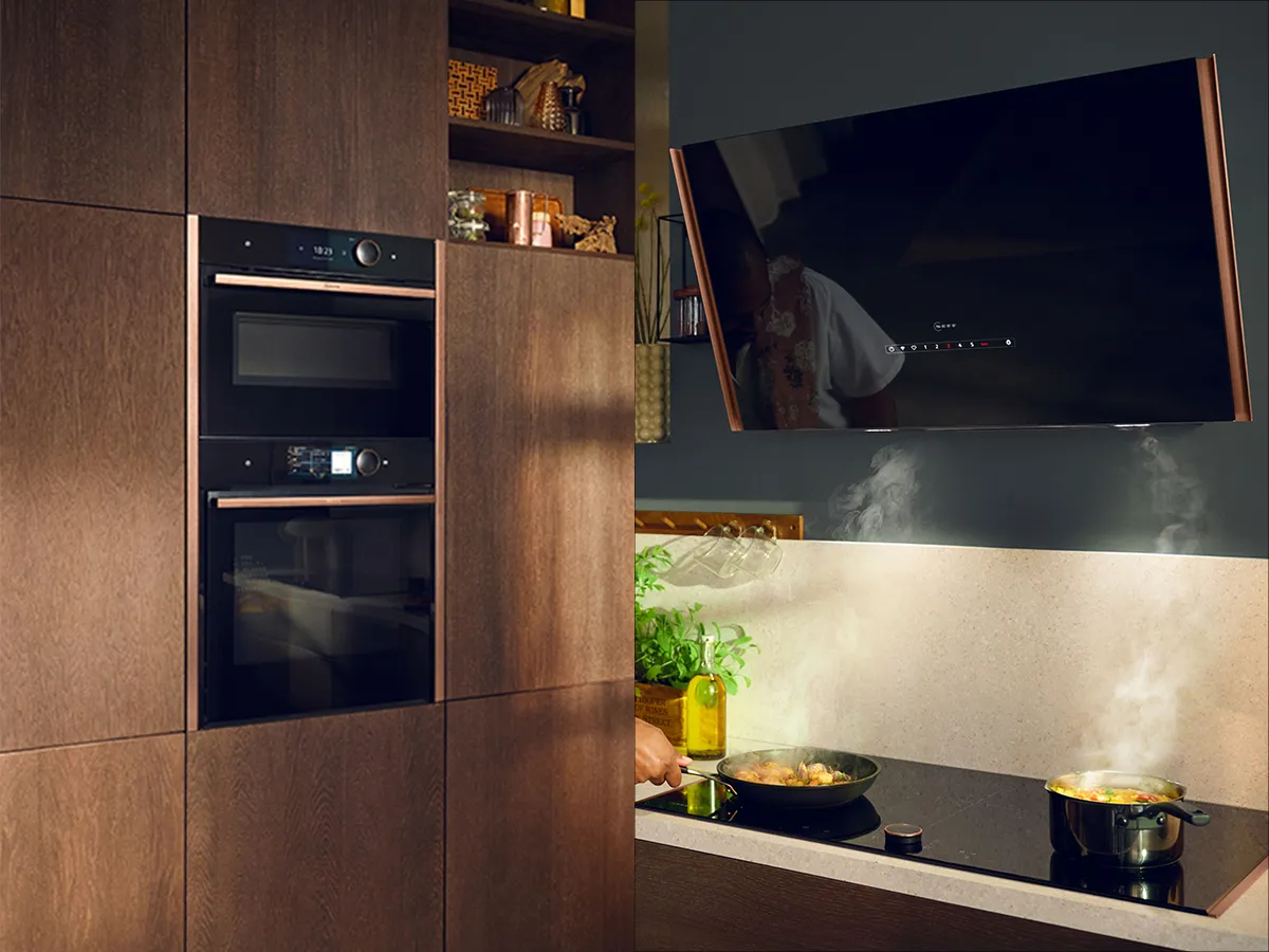 A modern kitchen with integrated black Neff appliances, including a microwave, oven, and a hob with cooking pots, all set into dark wooden cabinetry, with a large screen displaying a video above the hob.