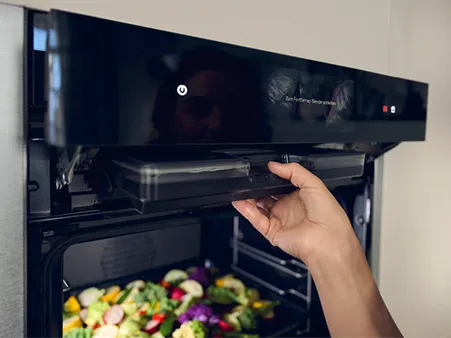 Eine Hand öffnet einen NEFF Ofen mit Dampfunterstützung, gefüllt mit buntem Gemüse.
