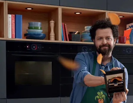 גבר לבוש בסווטשירט עם סינר עובד במטבח ומניח פירות או ירקות על מכשיר חיתוך, עם ארונות מודרניים ברקע.