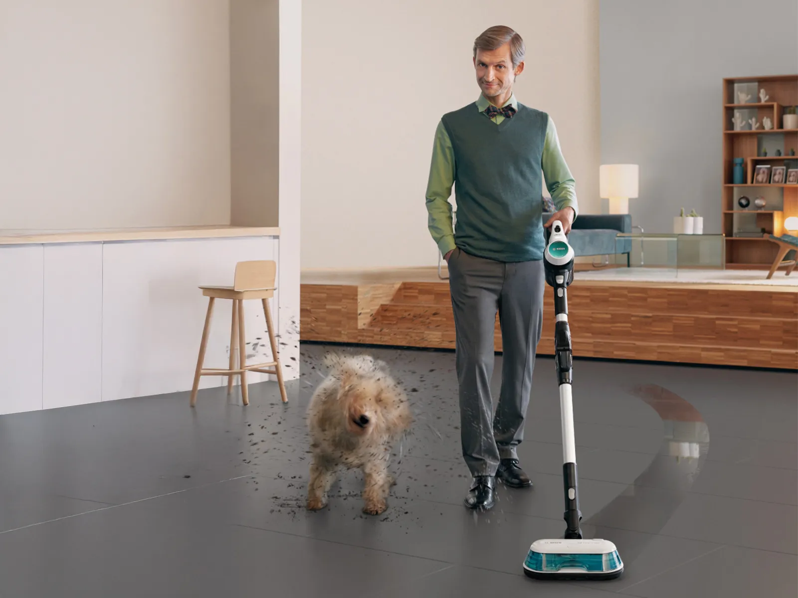 En välklädd man står och håller en dammsugare i ett modernt vardagsrum, medan en hund skakar av sig smuts i närheten.