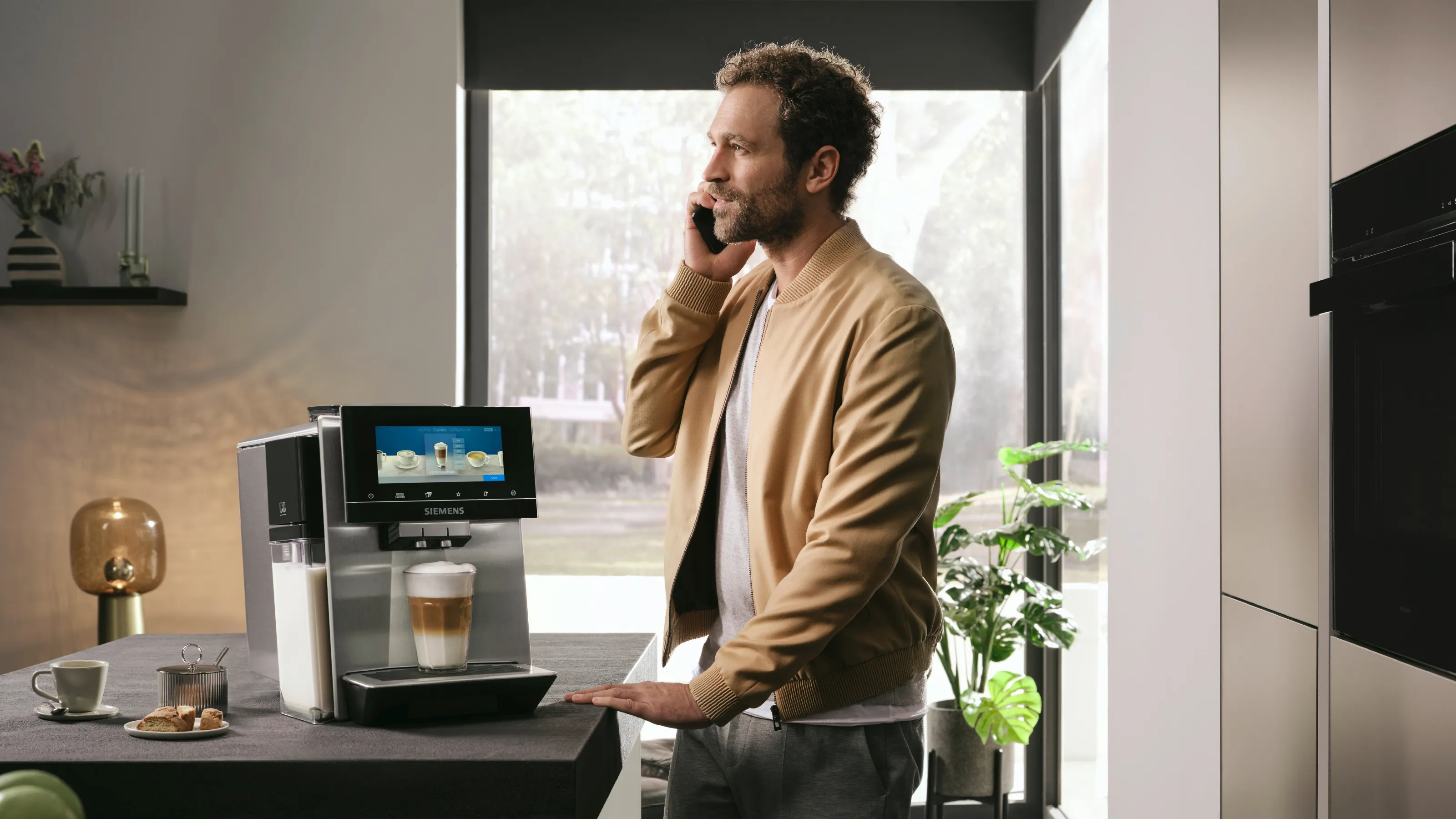 O persoană stă lângă o cafetieră, vorbind la telefon, cu un latte în față, înconjurată de o bucătărie modernă și plante.