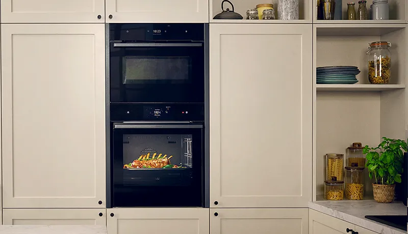 A seamless combination of two black NEFF built-in ovens is shown integrated into off-white, shaker-style kitchen cabinets. The bottom oven is illuminated, showing a rack of lamb cooking, while the top combination oven's display is also lit. To the right, open shelving displays kitchenware and food items.