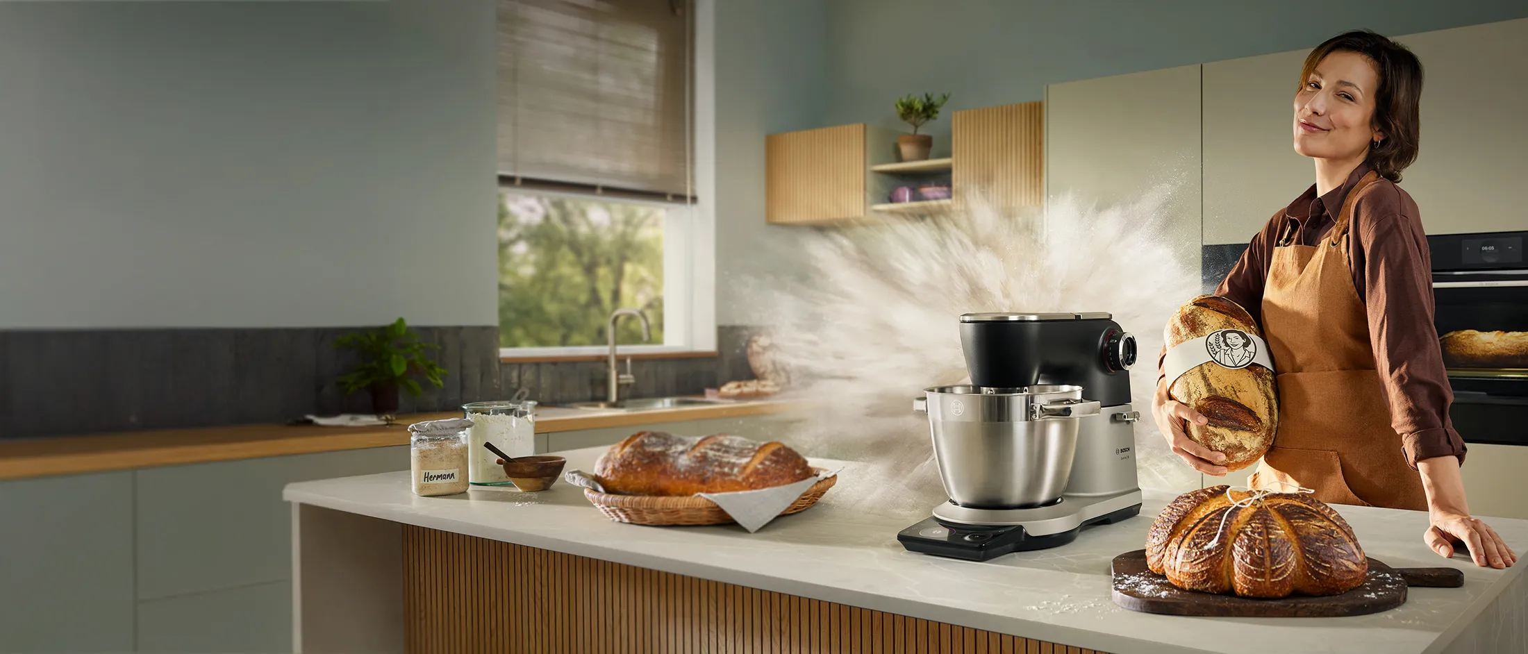 Frau in der Küche mit Bosch Mixer und frisch gebackenem Brot auf der Arbeitsplatte, umgeben von Zutaten.