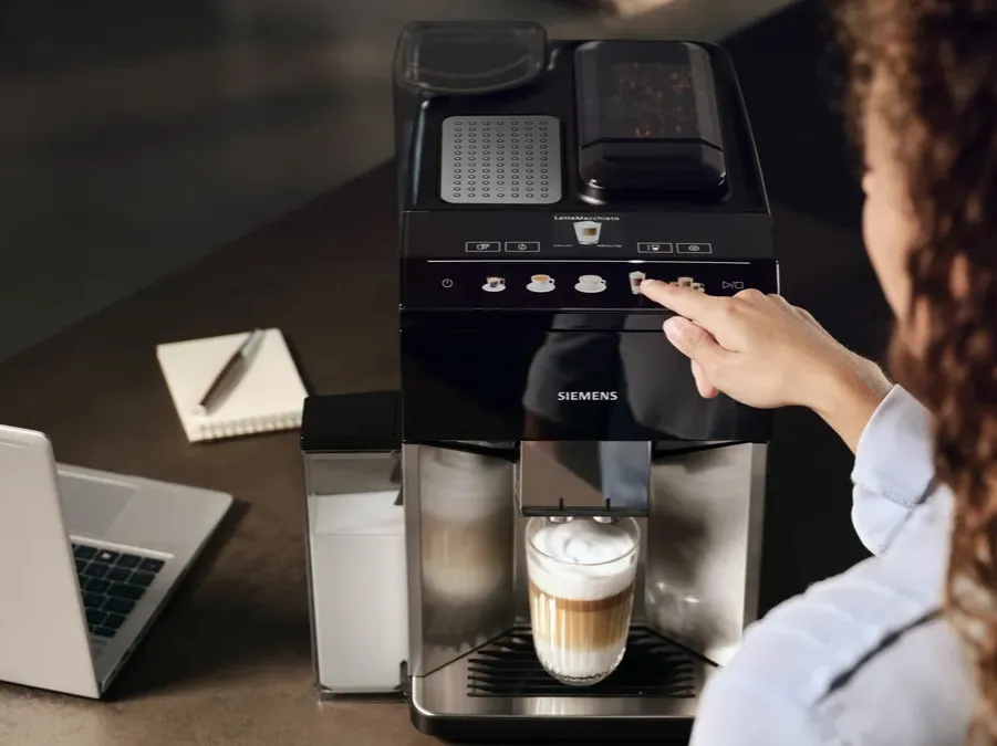 Woman is pressing the cappuccino button at the EQ500 coffee machine, a glass with coffee and milk foam already under the outlet.MASTER | Global | Coffee Machines - 