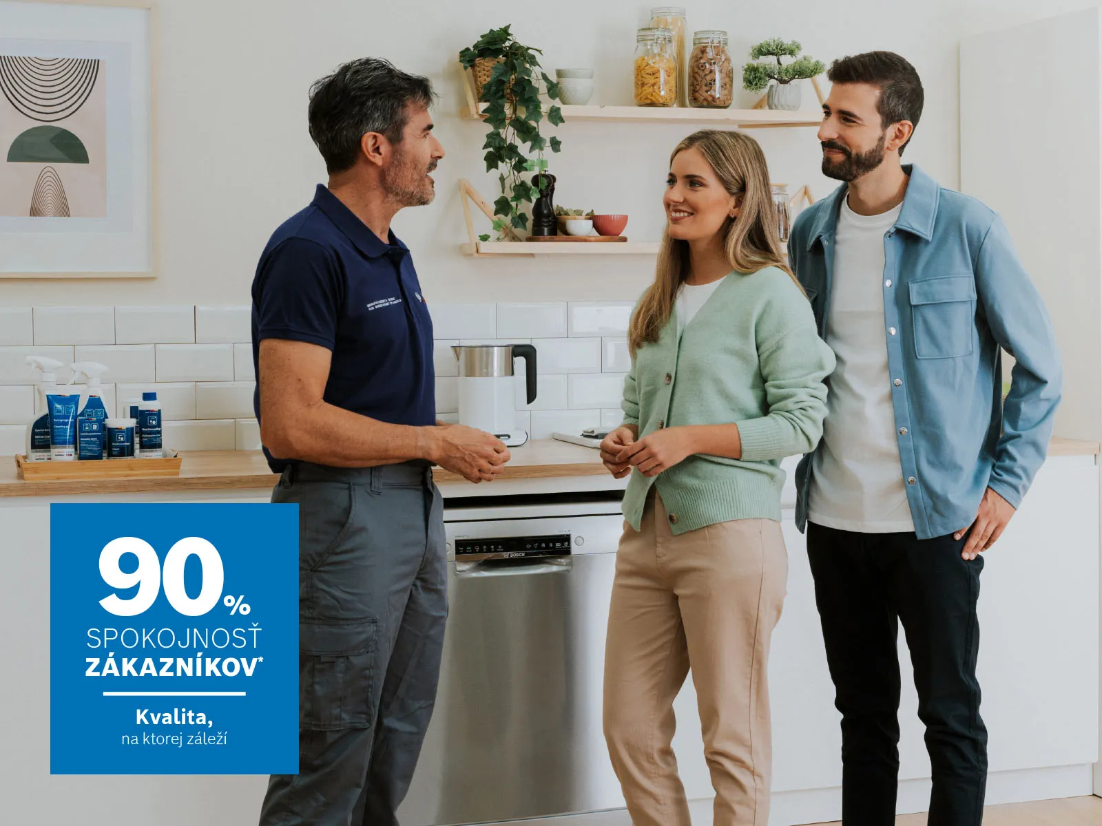 Servisný technik v tmavomodrej uniforme sa rozpráva s mladým párom v modernej svetlej kuchyni. Všetci sa usmievajú a stoja vedľa umývačky riadu