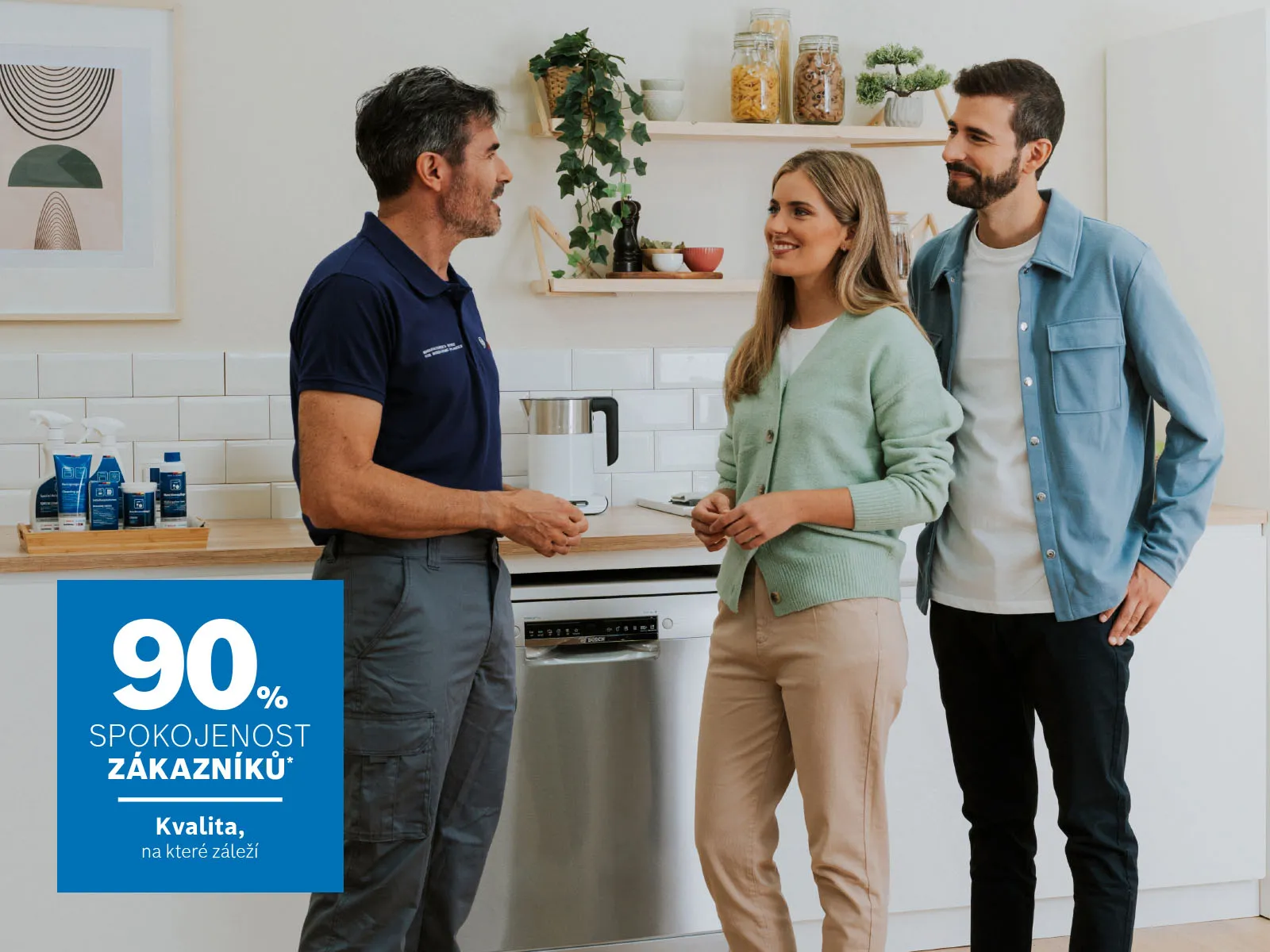 Servisní technik v tmavě modré uniformě hovoří s mladým párem v moderní světlé kuchyni. Všichni se usmívají a stojí vedle myčky nádobí.