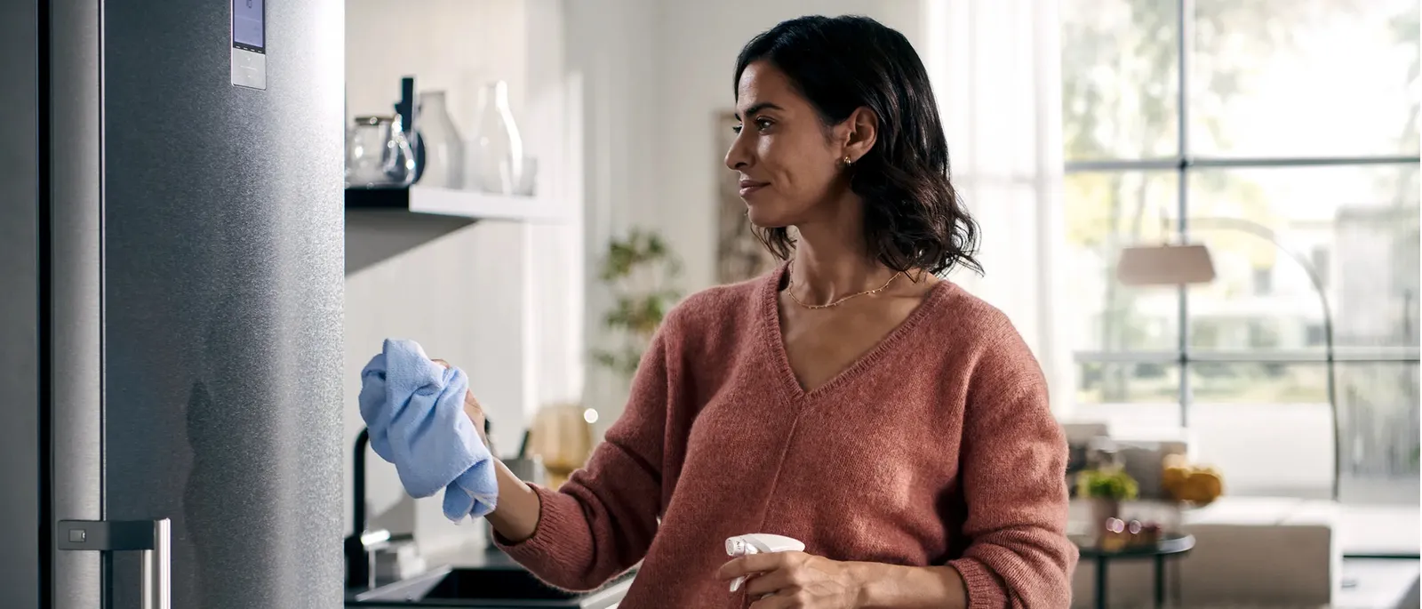 Una donna pulisce un frigorifero Siemens con un panno mentre tiene in una mano una bottiglia spray di detersivo.