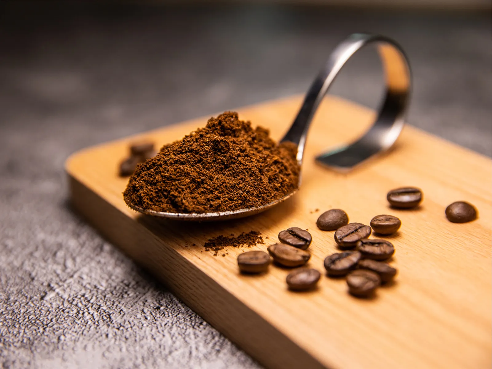 A spoonful of ground coffee rests on a wooden cutting board, surrounded by whole coffee beans. The background features a textured surface, adding depth to the image.