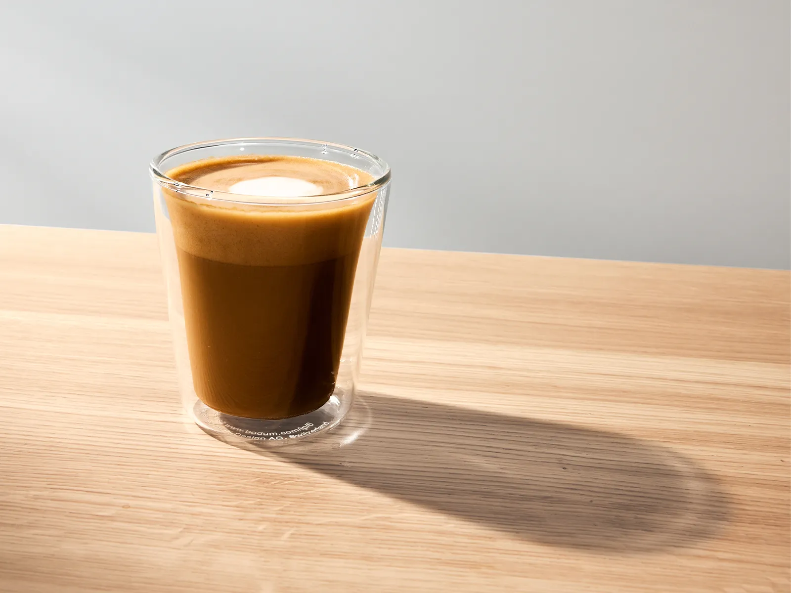 A clear glass cup filled with a creamy coffee beverage sits on a wooden surface, featuring a layer of frothed milk on top. Soft lighting highlights the cup and casts a gentle shadow beside it.