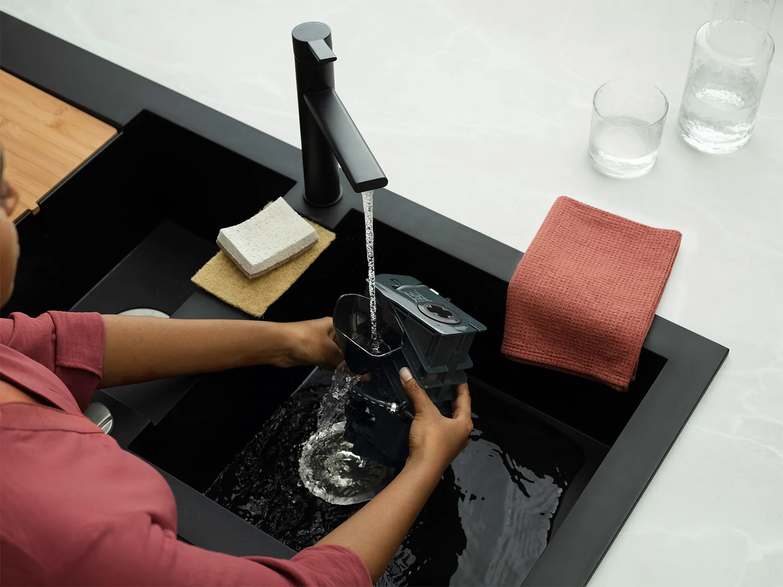 An overhead view of a kitchen sink with water flowing from a modern black faucet. A person is rinsing a dark-colored coffee maker part in the sink, while various cleaning items, including a sponge and a cloth, rest nearby. Two clear glasses are also visible on the countertop.