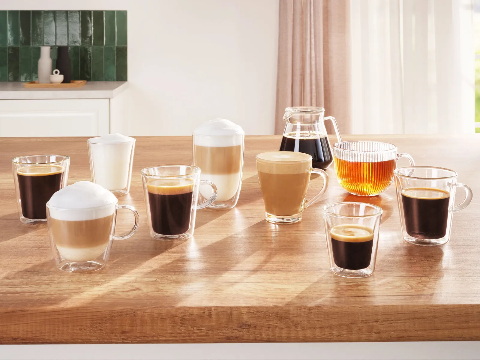 A wooden table features various glass coffee cups with lattes, espressos, and black coffee, alongside a carafe of brewed coffee and a striped pitcher of tea, all illuminated by sunlight in a cozy kitchen.