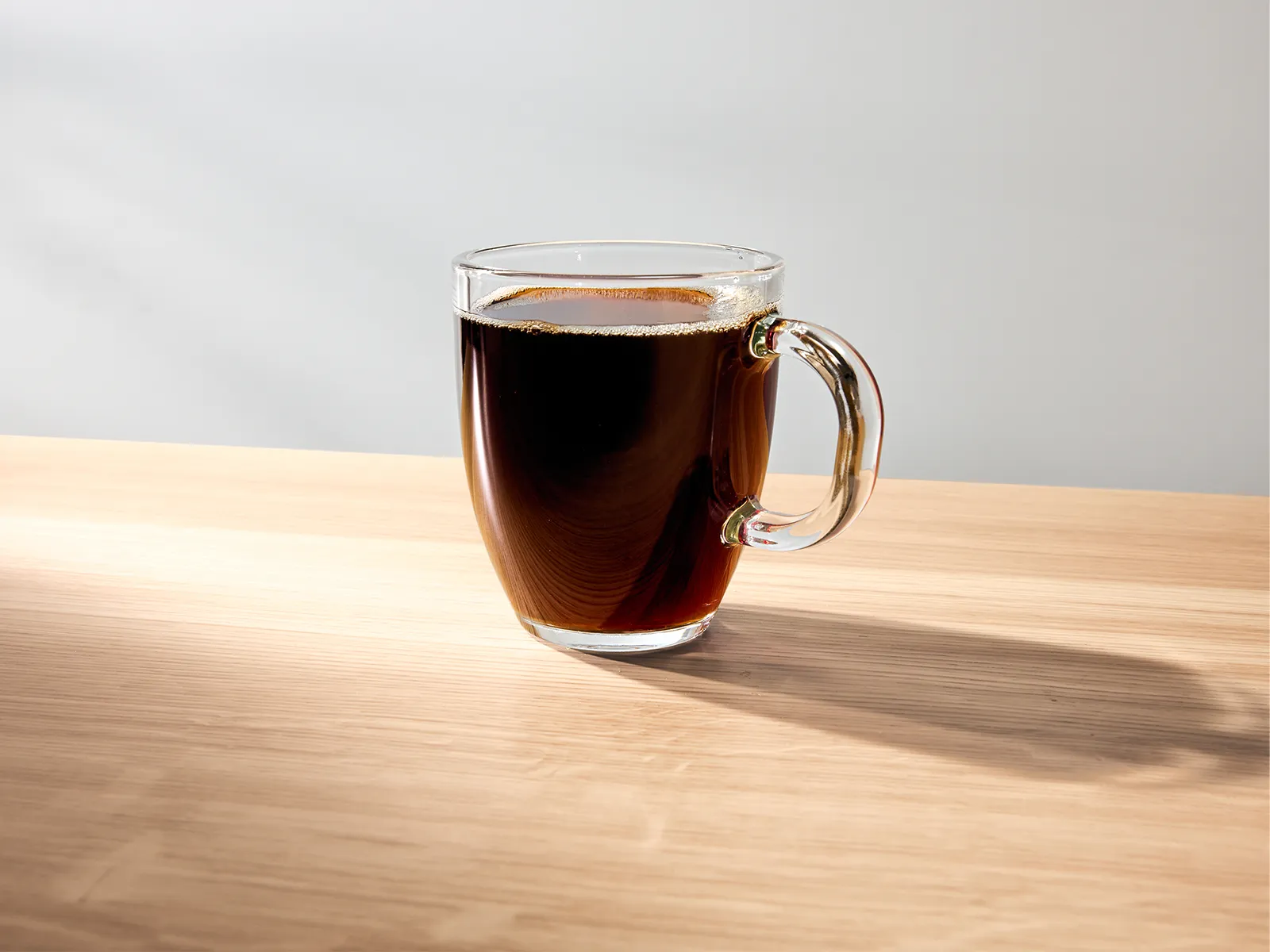 A clear glass cup filled with dark coffee sits on a wooden surface, illuminated by soft light that casts a gentle shadow beside it.