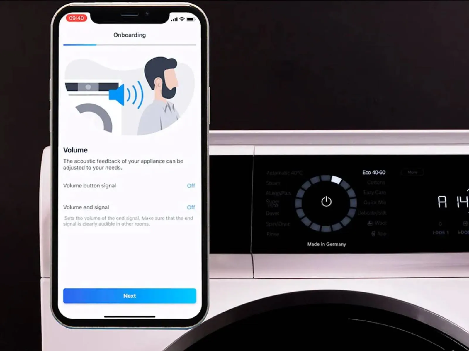 Išmanusis telefonas, rodantis prietaiso nustatymus, šalia modernios skalbimo mašinos su skaitmeniniu sąsaja ir valdymo ratuku.