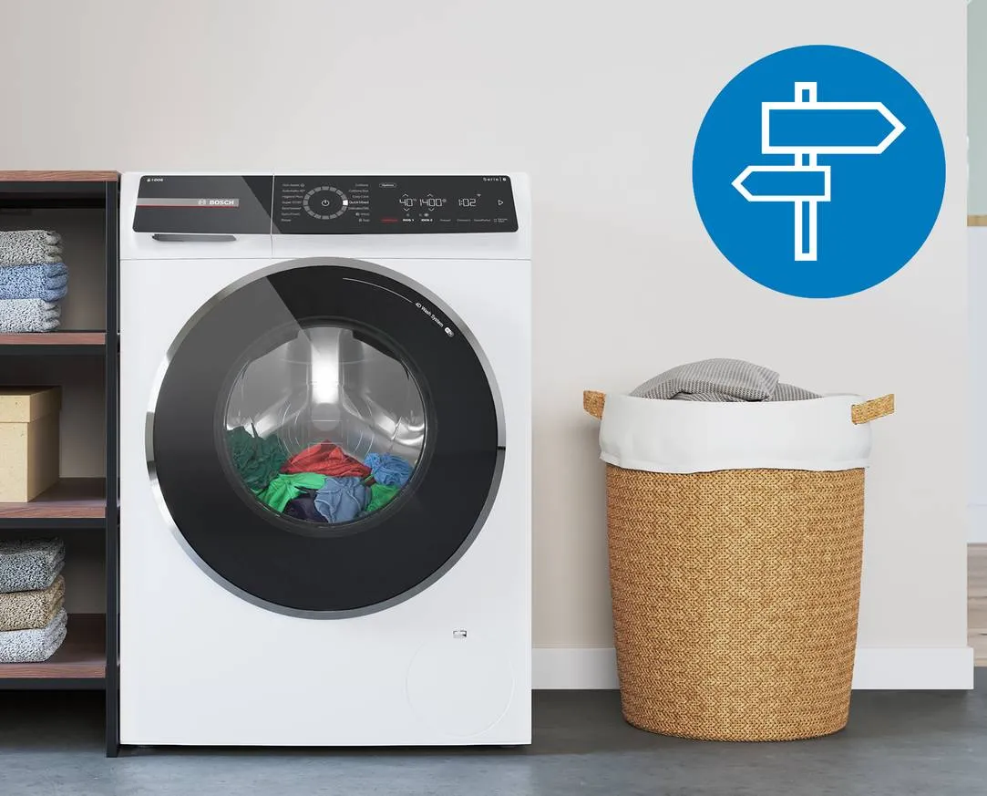 Moderne witte wasmachine met een doorzichtige deur, naast een open kast met opgevouwen handdoeken en een houten doos. Naast de wasmachine staat een rieten wasmand. In de rechterbovenhoek is een blauw pictogram met een richtinggevende teken.