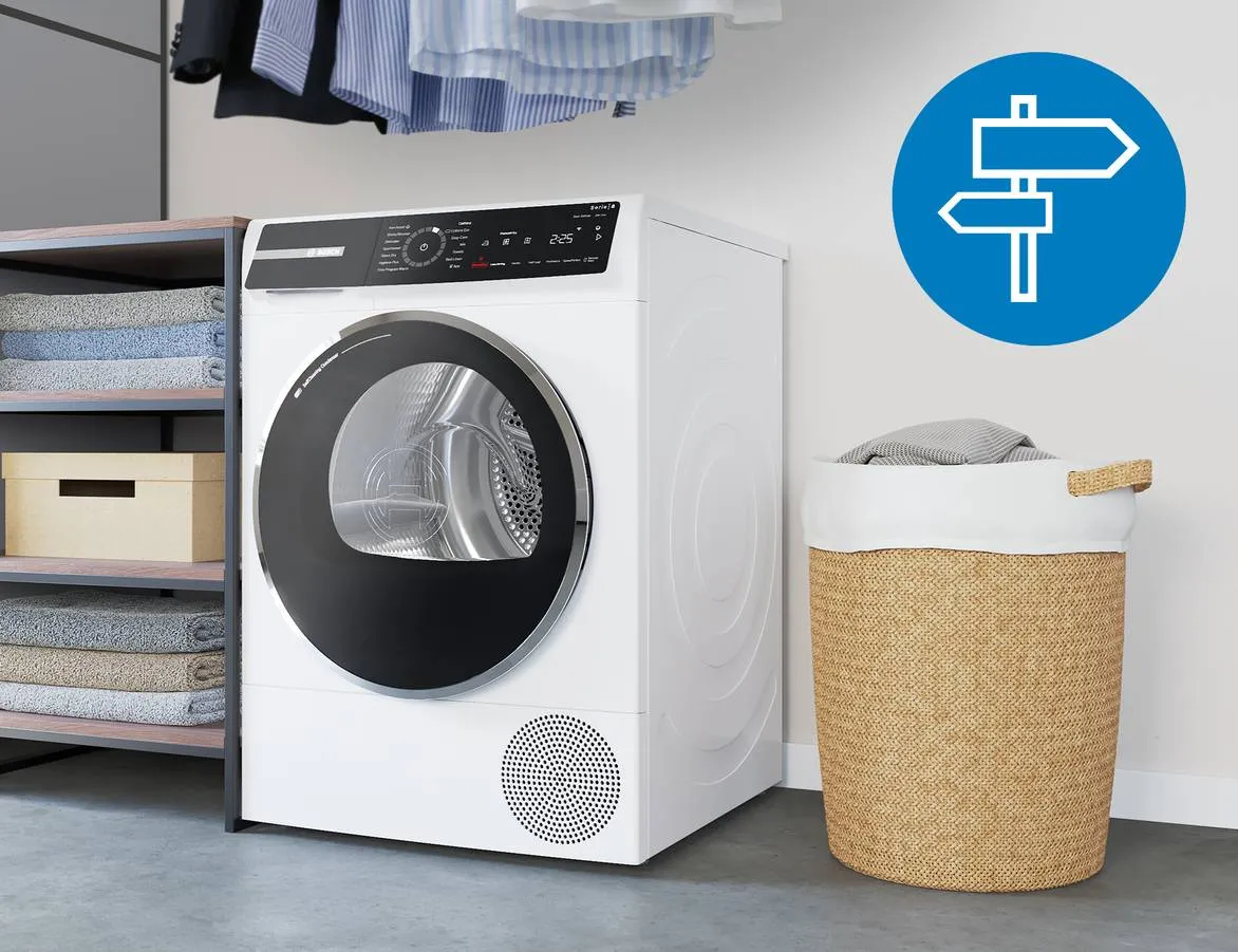 Moderne witte wasmachine met een zwarte deur, naast een open kast met opgevouwen handdoeken, een houten doos en een rieten wasmand met een witte rand. Boven de wasmachine hangen shirts en in de rechterbovenhoek is een blauw pictogram met richtinggevende symbolen