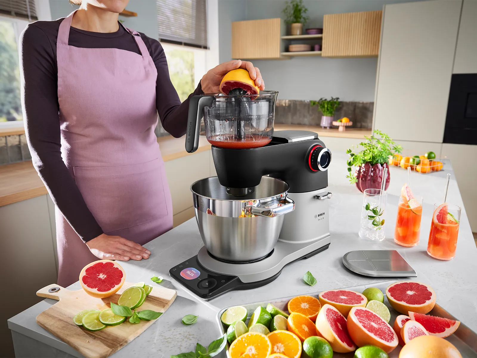The Series 8 Stand Mixer stands on a kitchen counter with various fruits and juices around it, while a person uses the citrus press accessory on the mixer to make juice.
