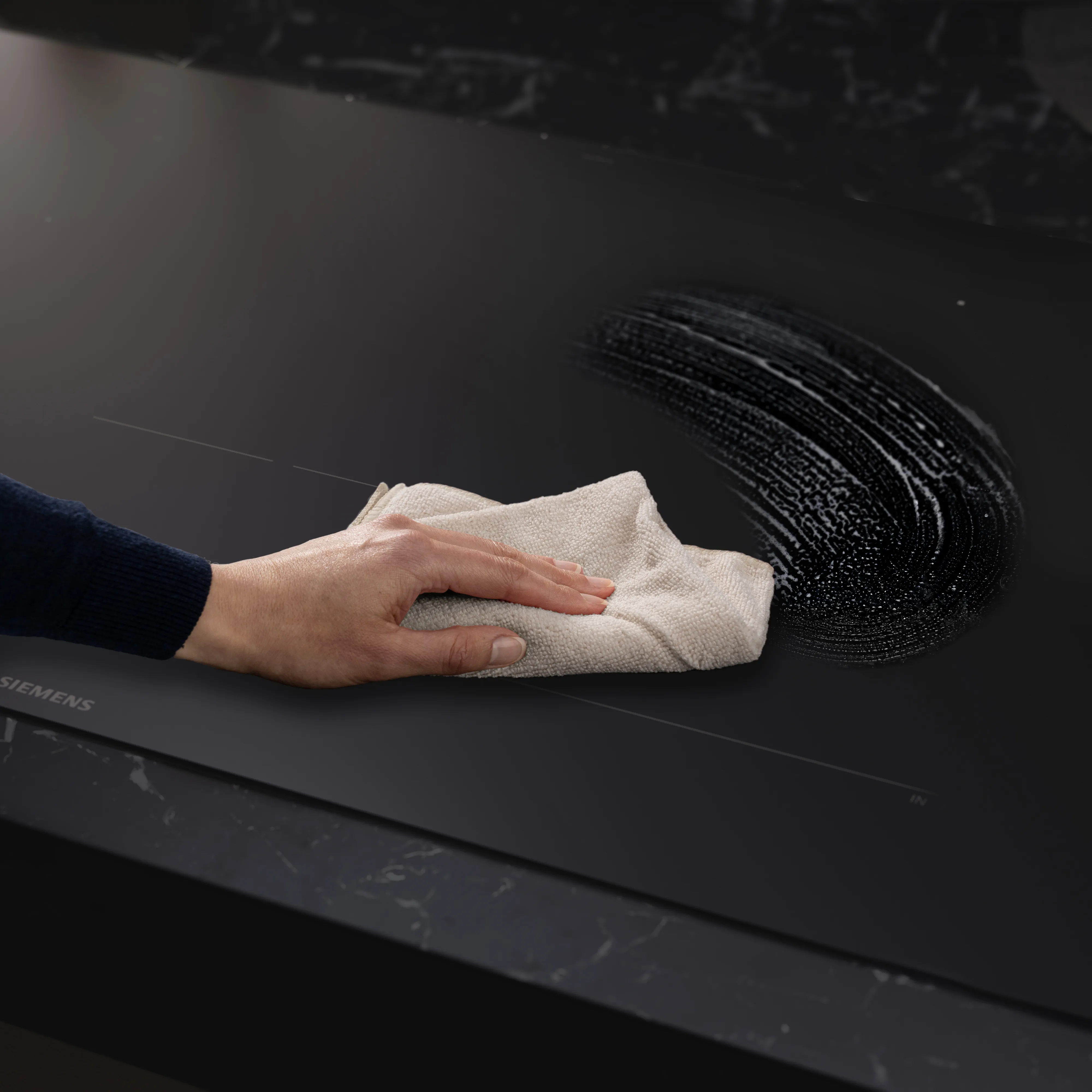 Hand wiping a black glass stovetop with a beige cloth, removing a semi-circular area of grease or residue.