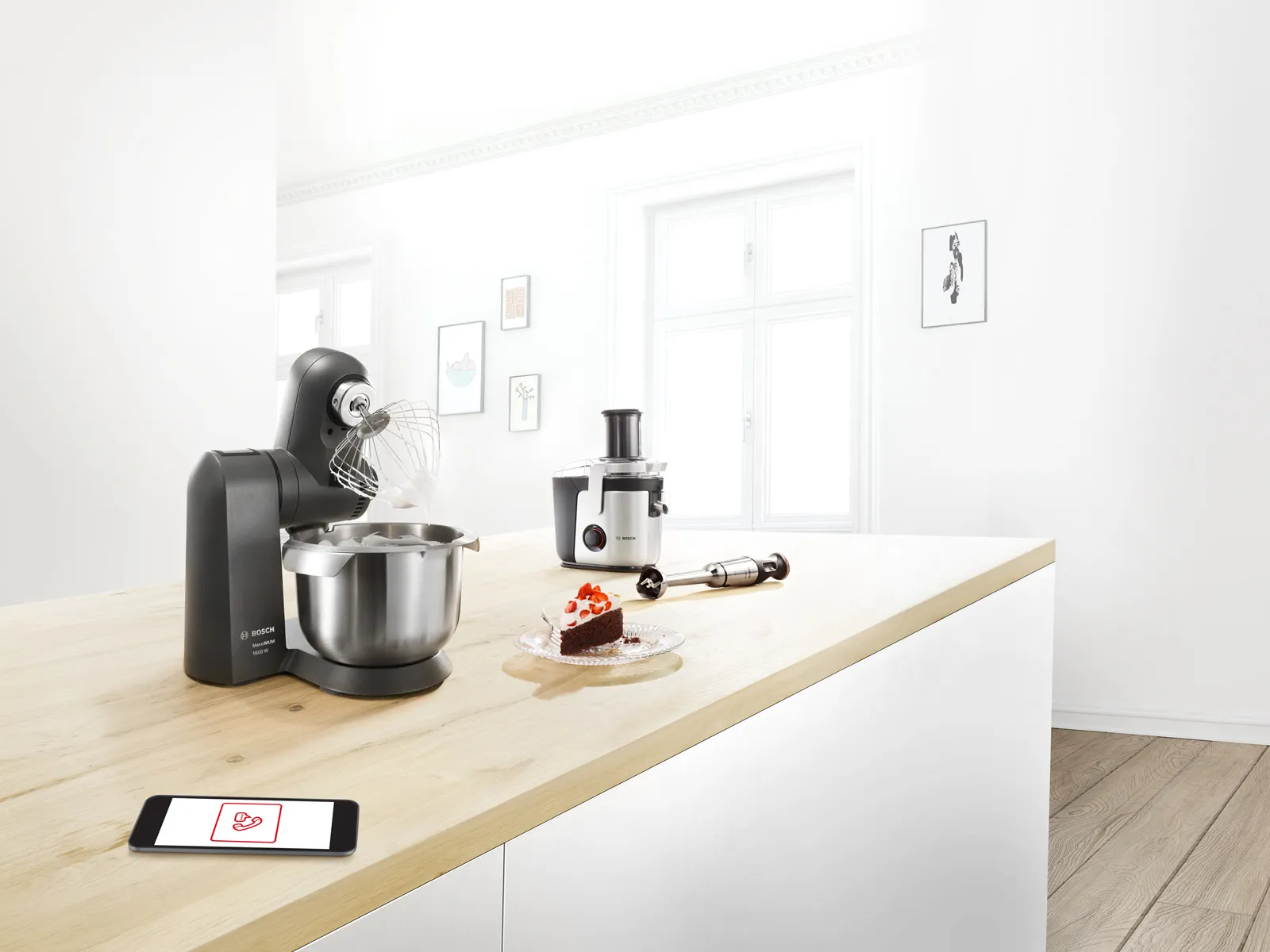 מיקסר של Bosch במטבח מודרני, קערה מתכתית עם קצפת, עוגה ושולחן עם סמארטפון. אווירה מוארת ונקייה.