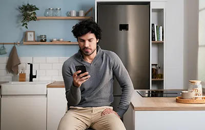 alt-Un hombre zit op een keukenteller en kijkt naar zijn smartphone, op zoek naar tips voor het oplossen van foutcodes, met planken en een koelkast op de achtergrond.