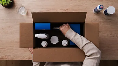 alt-Een persoon reikt in een bruine kartonnen doos vol met verschillende schoonmaak- en verzorgingsproducten, inclusief flessen en verpakkingen, die op een houten tafel staat.