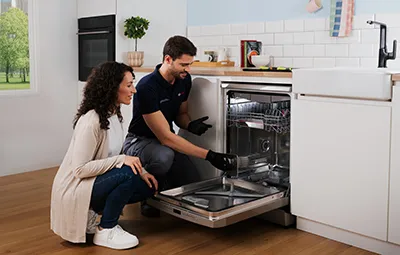 alt-Een technicus verleent een reparatiedienst terwijl hij de werking van de vaatwasser demonstreert aan een vrouw in een moderne keuken, waarbij hij de functies en het onderhoud benadrukt.