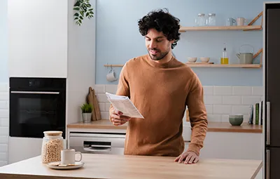 alt-Een man leest een gebruiksaanwijzing in een gezellige keuken, terwijl hij een bruine trui draagt. De keuken heeft houten planken, planten en een pot met ontbijtgranen op het aanrecht.
