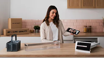 alt-Een vrouw glimlacht terwijl ze reserveonderdelen vasthoudt voor een keukenapparaat in een moderne keuken met houten kasten en een schoon aanrecht.