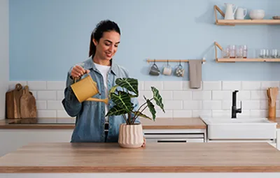 alt-Osoba podlewa doniczkową roślinę żółtym konewką na drewnianym blacie kuchennym, otoczoną dekoracjami domowymi, podkreślając ochronę gwarancyjną produktu.