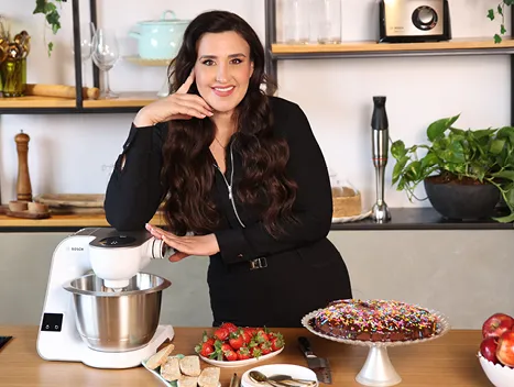 אישה עומדת ליד מכונת מיקסר, עם פירות ועוגה צבעונית על השולחן, בסביבה ביתית מזמינה.