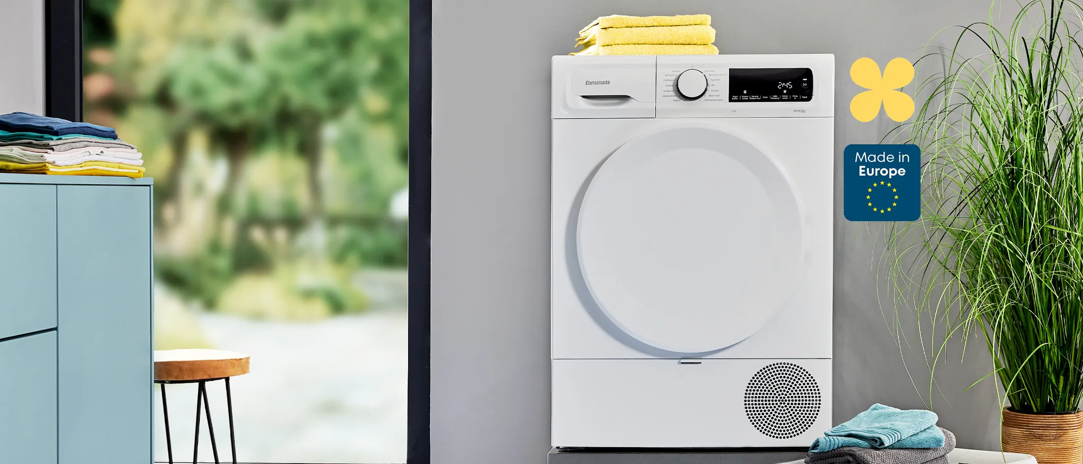 Ein weißer Trockner mit gefalteten Handtüchern steht in einer modernen Waschküche neben grünen Pflanzen und bunt gestapelter Wäsche mit der Aufschrift „Made in Europe“.