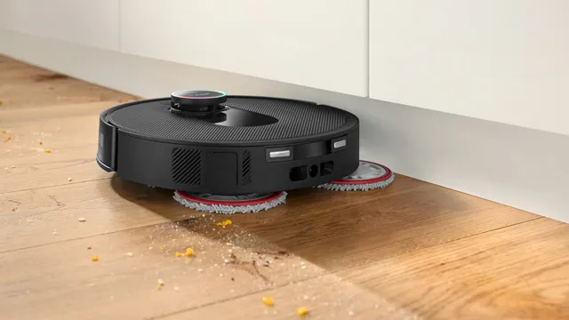 Ein schwarzer Saugroboter mit Wischtüchern reinigt den Boden unter einem Küchenschrank, umgeben von Krümeln.