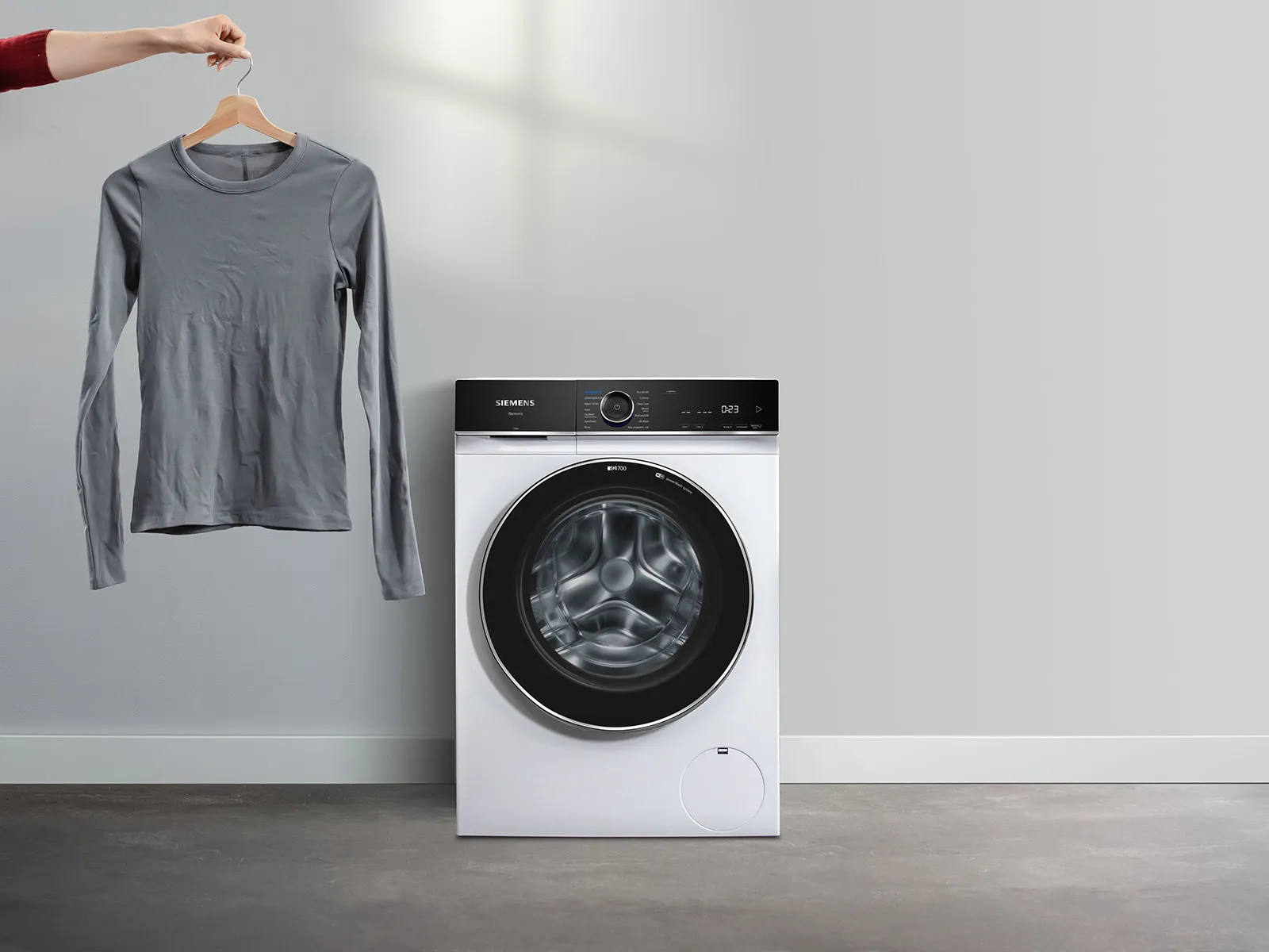 Die Hand einer Person hält ein graues Langarmhemd über einer eleganten, modernen Siemens-Waschmaschine in einem minimalistischen Waschraum.
