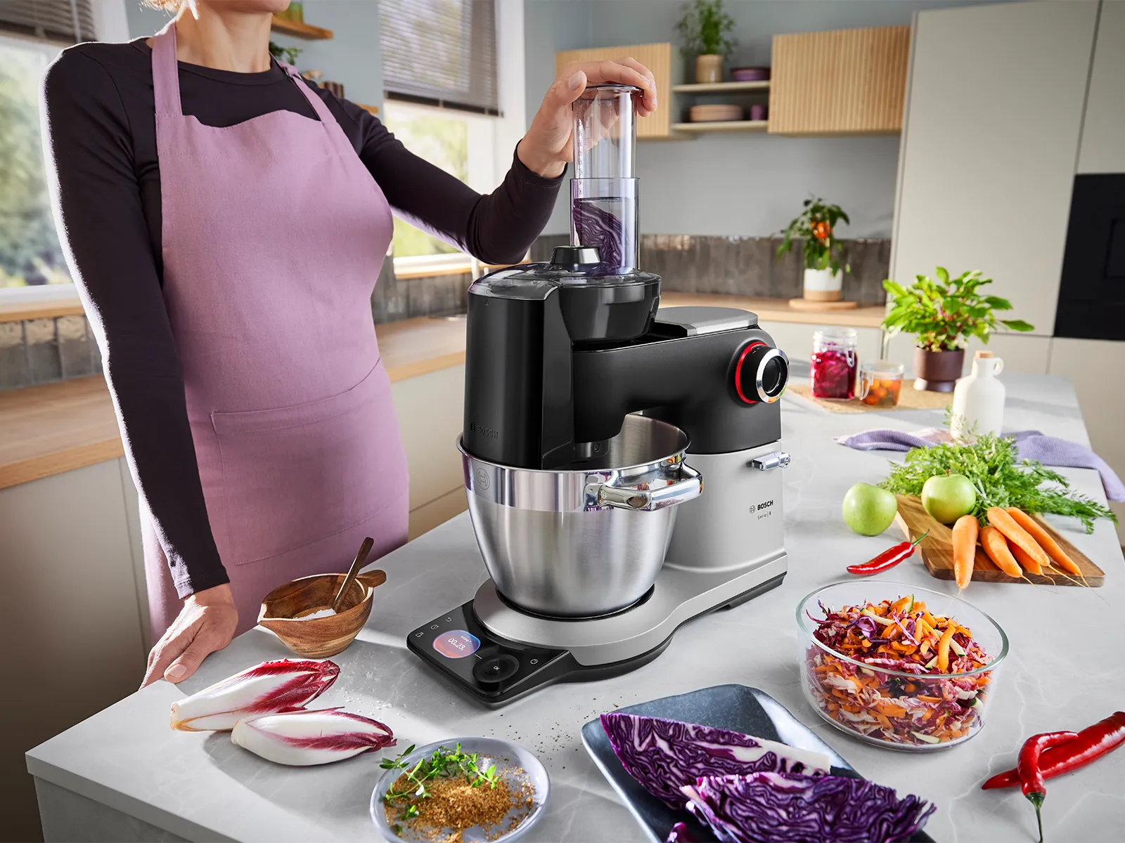 Le robot pâtissier de la Série 8 est posé sur un plan de travail de cuisine, entouré de divers fruits et légumes coupés, tandis qu'une personne utilise l'accessoire râpeur.