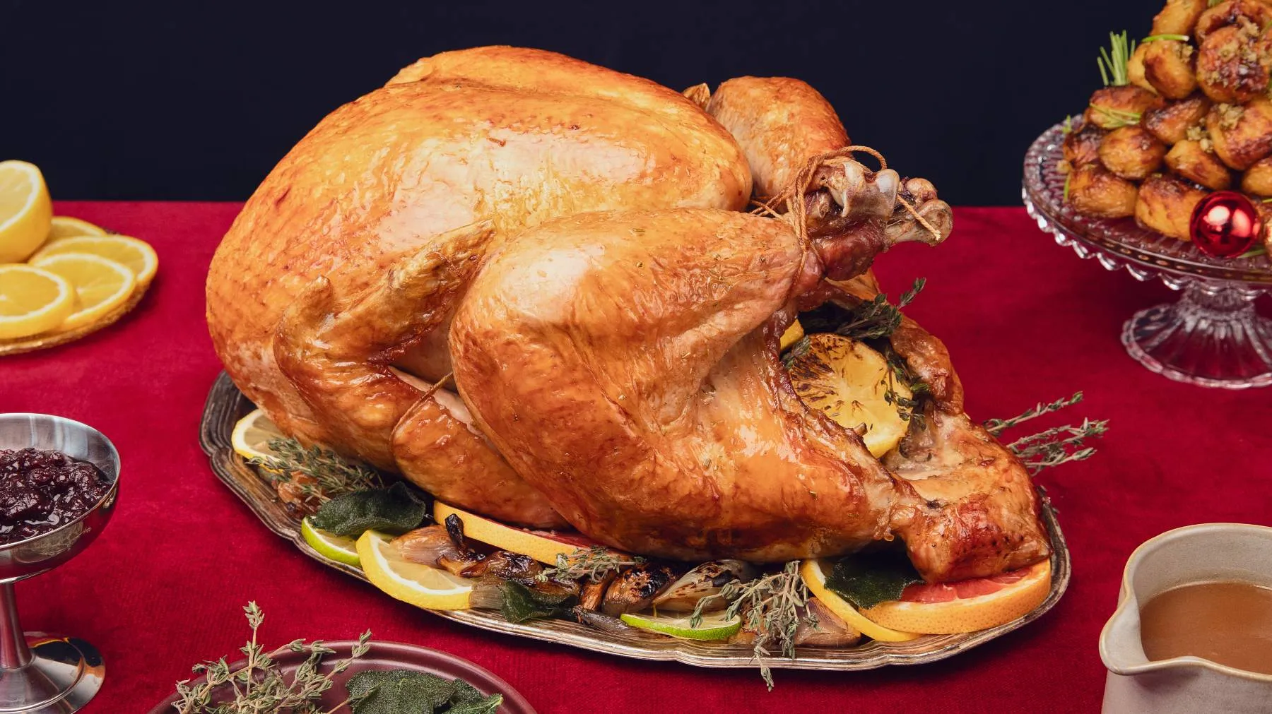 A perfectly roasted golden-brown turkey, garnished with lemons, grapefruit, and herbs, on a silver platter on a red tablecloth, surrounded by cranberry sauce, gravy, and roasted potatoes.