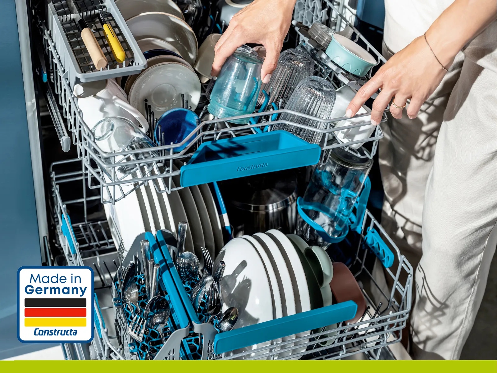 Hände räumen Geschirr und Gläser in einen geöffneten Constructa Geschirrspüler ein. "Made in Germany"-Label.