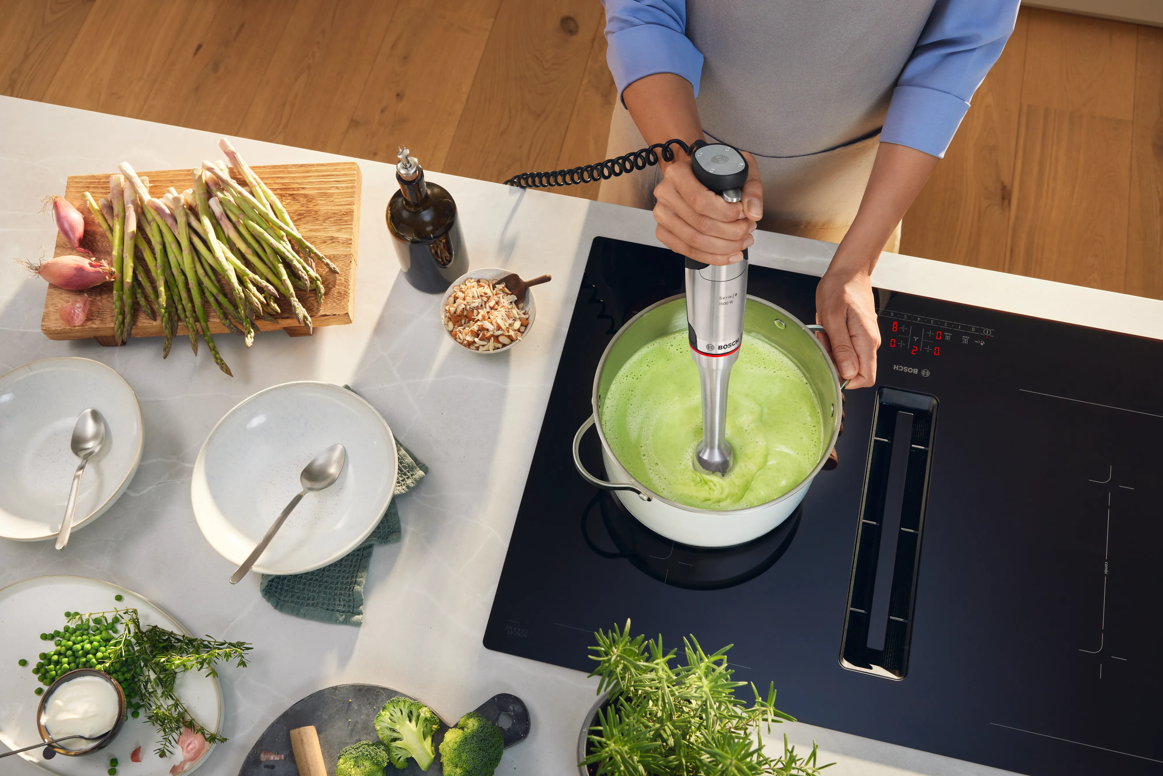 Person blending green soup in pot on stovetop surrounded by fresh vegetables and herbs on kitchen counter.