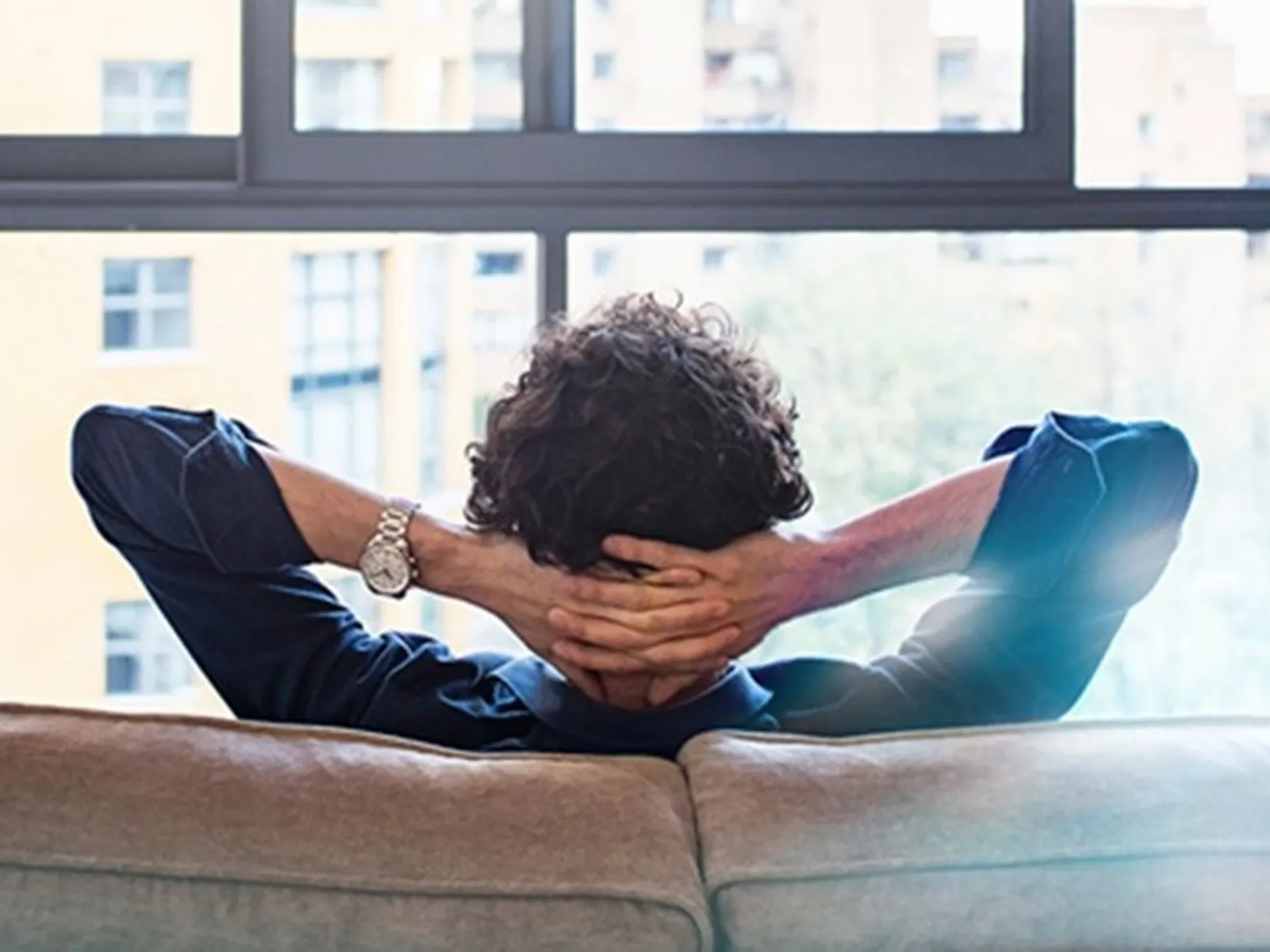 Ένας άνθρωπος όπου κάθεται σε έναν καναπέ και αγναντεύει χαλαρός την θέα του από την τζαμαρία του σπιτιού του.