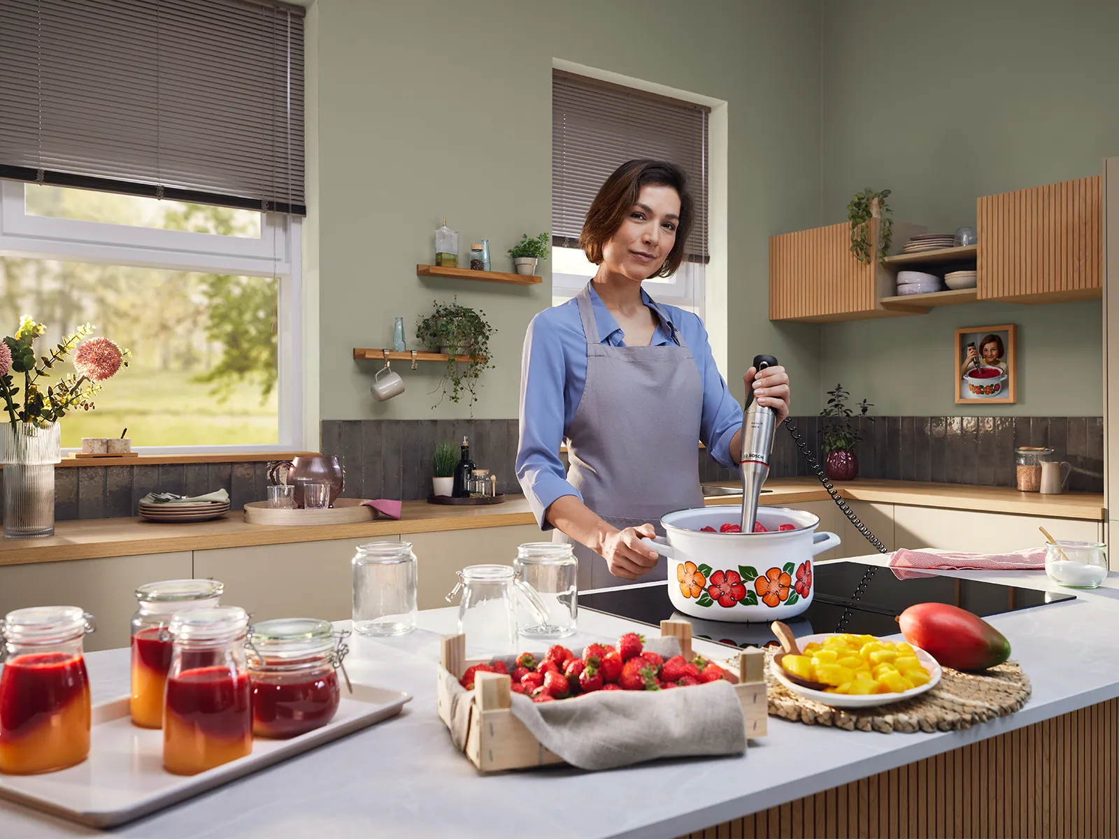 Una mujer está mezclando en una vieja sartén con el Ergomaster Serie 8, mientras que a su alrededor hay varias frutas y mermeladas en la encimera de la cocina.