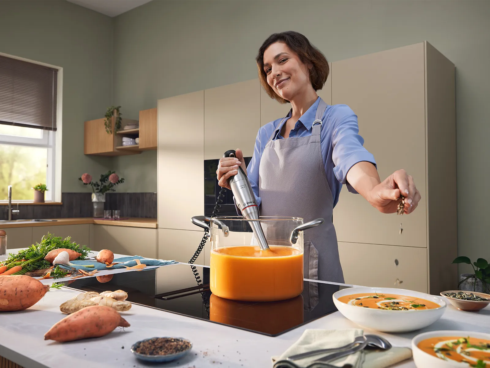 Una mujer está mezclando con una mano en una cacerola de vidrio con el Ergomaster Serie 8, mientras agrega ingredientes a una sopa con la otra mano.